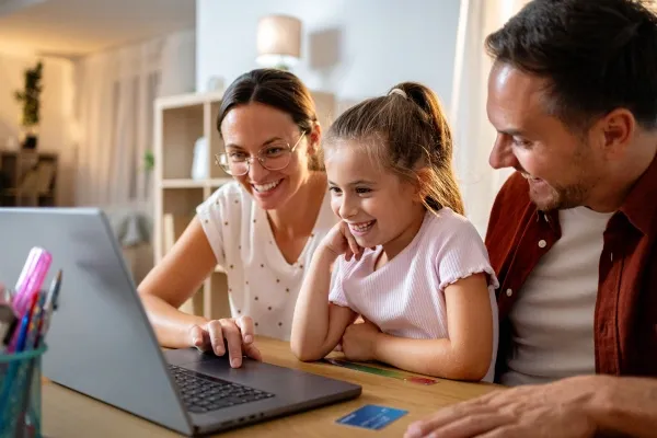 A smiling family, consisting of a mother, father, and young daughter, sits together at a table looking at a laptop screen, with a credit card visible on the table.